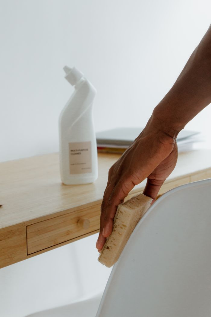 A minimalist room showing a person cleaning a chair with a sponge and a cleaning product.